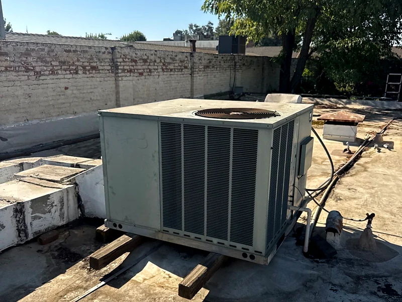 A rooftop package unit sitting on low wood blocks over a flat roof, with ductwork running beneath it
