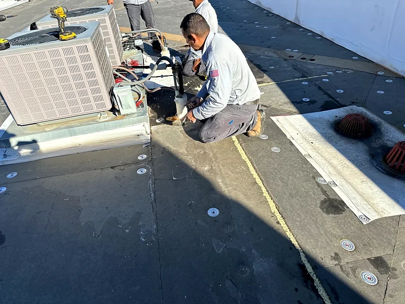 A Grandmark roofer sealing around a rooftop unit on a commercial flat roof, with insulation board and plates fastened across the deck