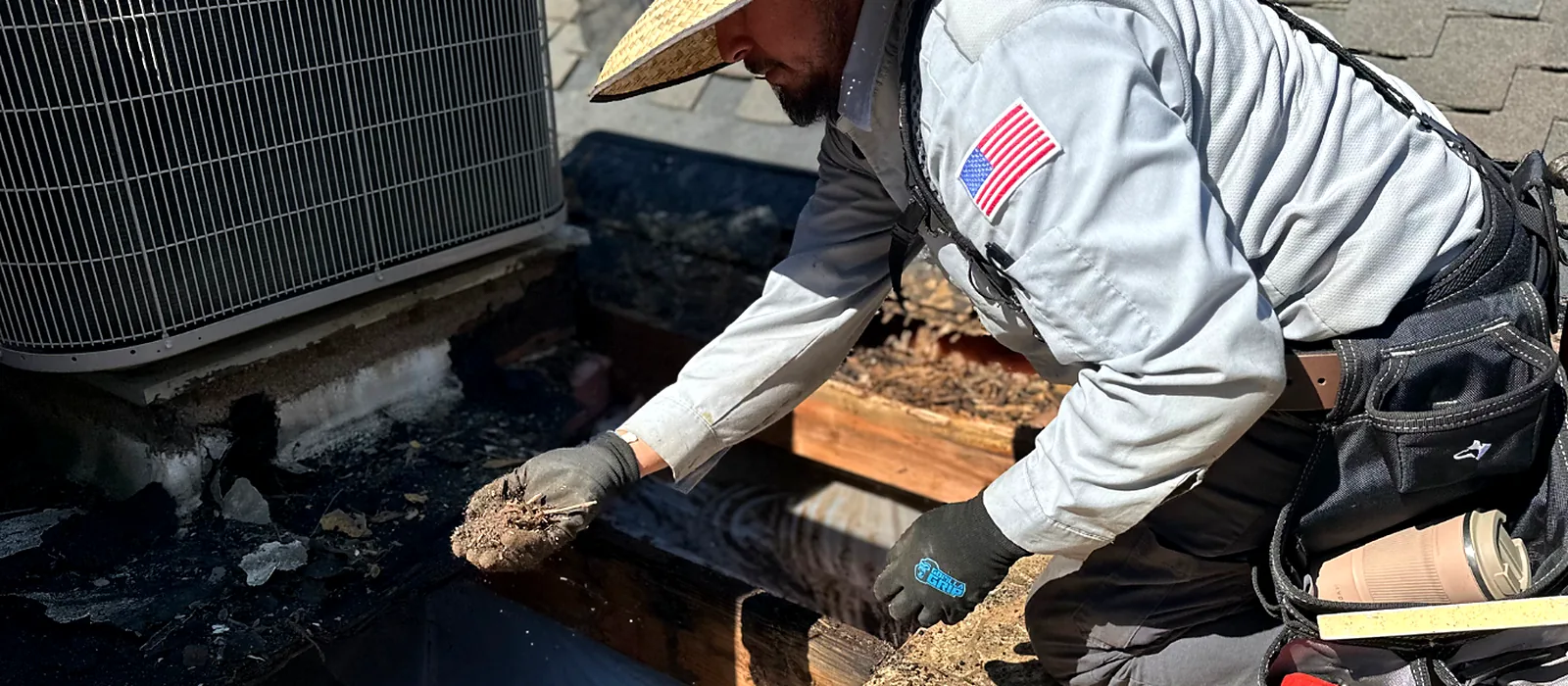 A Grandmark inspector pulling packed debris from a rotted curb beside a rooftop air conditioning unit during a second-opinion roof inspection