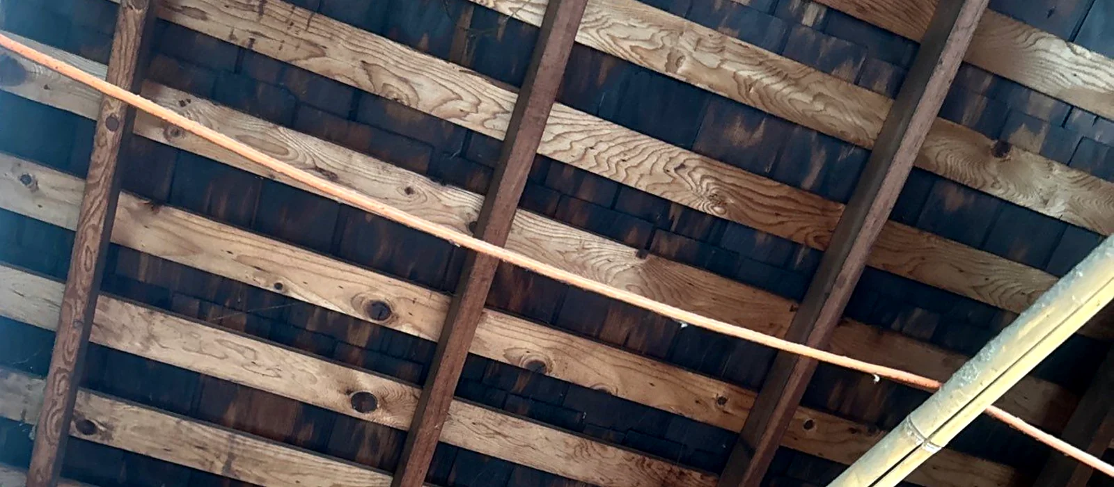 Underside of a wood shake roof seen from the attic, showing spaced skip sheathing with daylight and staining between the boards