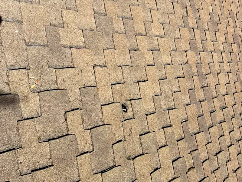A hole punched through a composition shingle on a Kerman roof, surrounded by shingles with heavy granule loss and debris caught in the courses